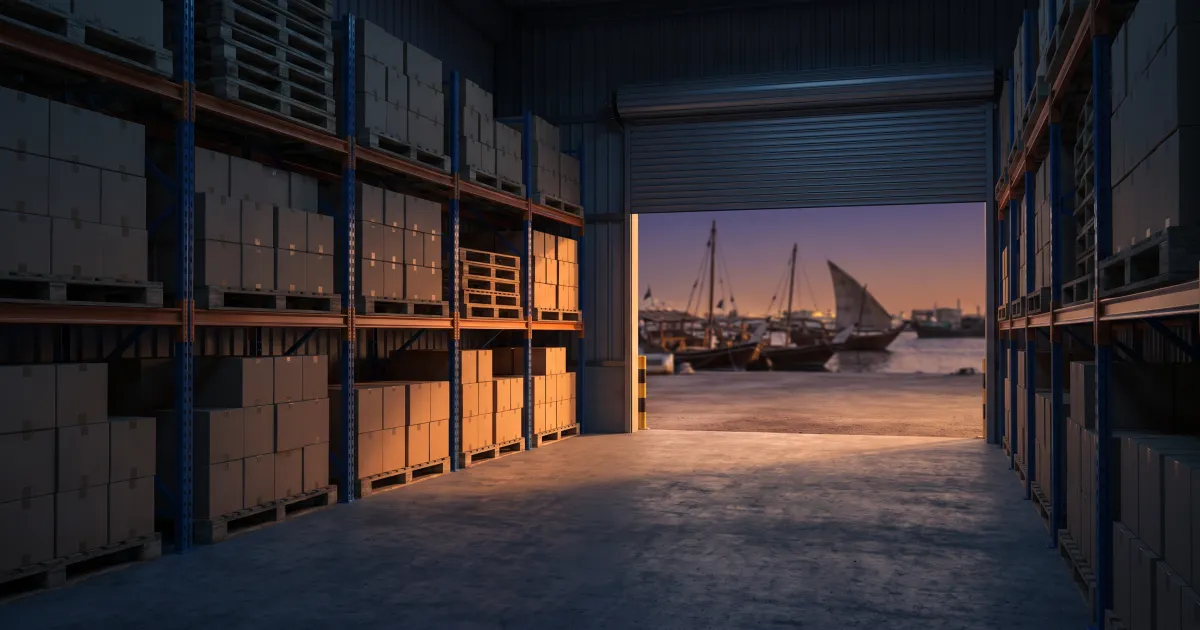 Plain cartons stacked on warehouse racking beside a half-open shutter overlooking wooden dhows moored on a Dubai creek at dusk