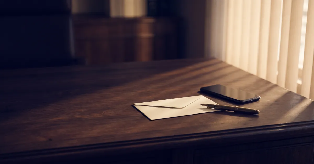 A sealed cream envelope beside a face-down smartphone and a fountain pen on a dark wooden desk in soft afternoon light