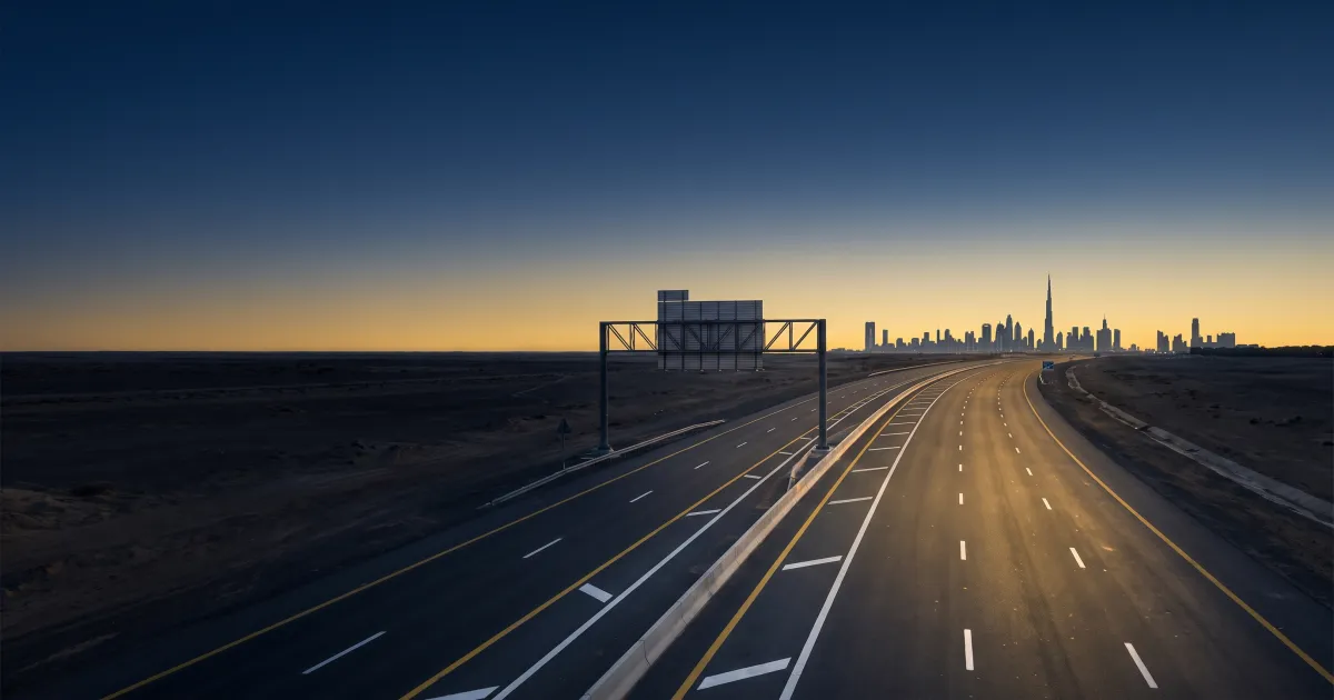 Empty multi-lane UAE highway at dusk with clear lane markings curving toward a distant city skyline under a deep blue sky